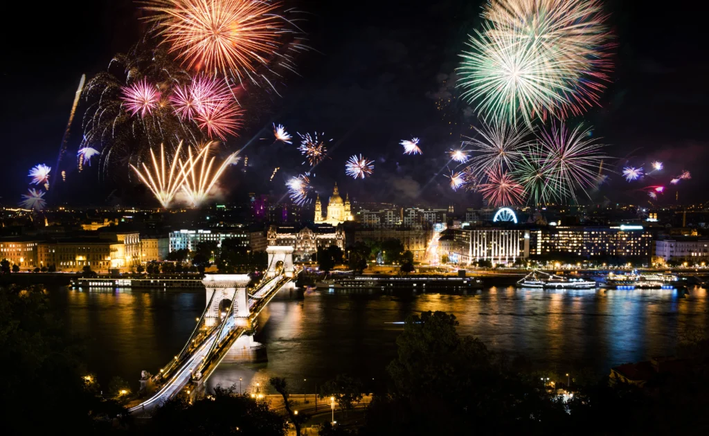 Colorful fireworks display over the illuminated Buda Castle complex in Budapest at night, with the grand baroque palace buildings lit in golden light reflected on the Danube River below.