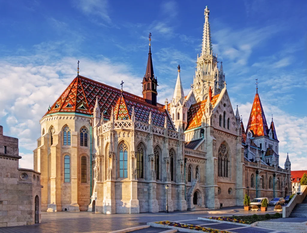 Matthias Church (Church of Our Lady) in Budapest's Castle District, featuring its distinctive colorful geometric tile roof and Gothic Revival architecture, with the medieval Fisherman's Bastion archway visible in the foreground.