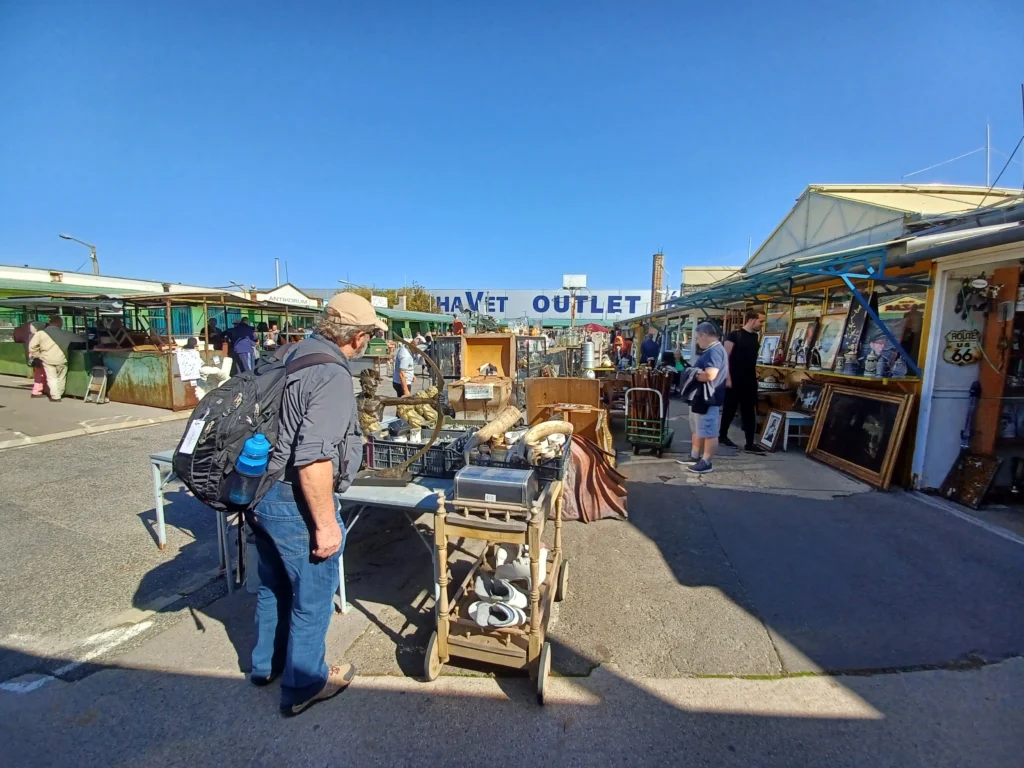 Shoppers browsing the outdoor stalls at Ecseri Flea Market in Budapest on a clear day, surrounded by a variety of antiques, vintage items, and collectibles displayed in an open-air setting.